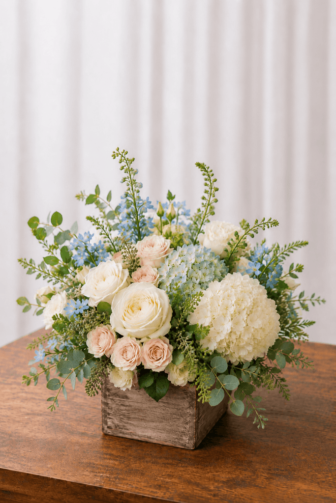 Coastal floral arrangement with ivory roses, white hydrangea, blush flowers, pale blue accents, and greenery in a square wooden box.