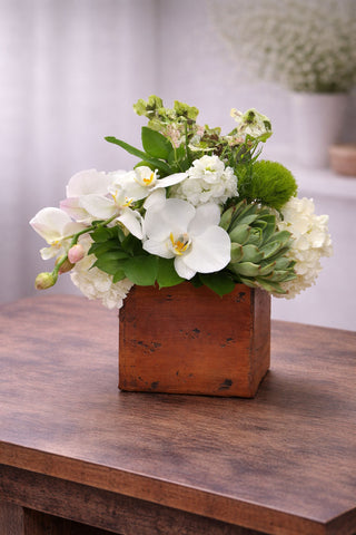 Penelope floral arrangement in a rustic wooden cube with white phalaenopsis orchids, hydrangea, succulents, green trick dianthus, and seasonal blooms.