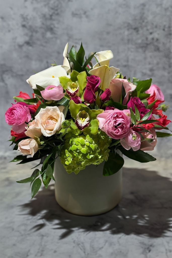 Vase arrangement featuring pink and blush roses, green hydrangea, cymbidium orchids, ranunculus, and calla lilies, styled in a modern ceramic container.