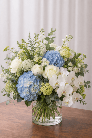 Coastal floral arrangement with blue hydrangeas, white roses, white orchids, airy white flowers, and greenery in a clear cylinder vase.