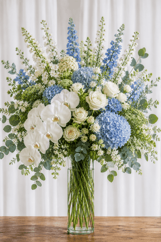 Coastal statement floral arrangement with blue hydrangeas, white orchids, ivory roses, airy white flowers, and greenery in a tall clear cylinder vase.