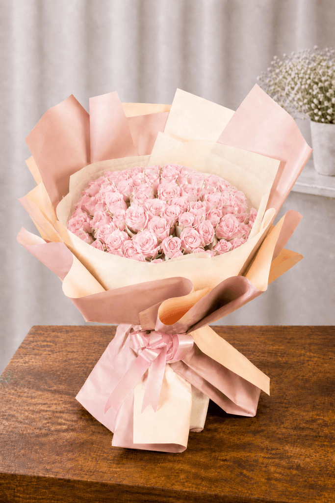 Hand-tied pink rose bouquet wrapped in blush and cream paper with a red ribbon, displayed on a wooden table in a soft studio setting.