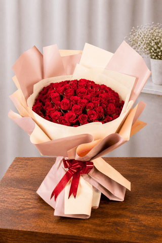 Hand-tied red rose bouquet wrapped in blush and cream paper with a red ribbon, displayed on a wooden table in a soft studio setting.