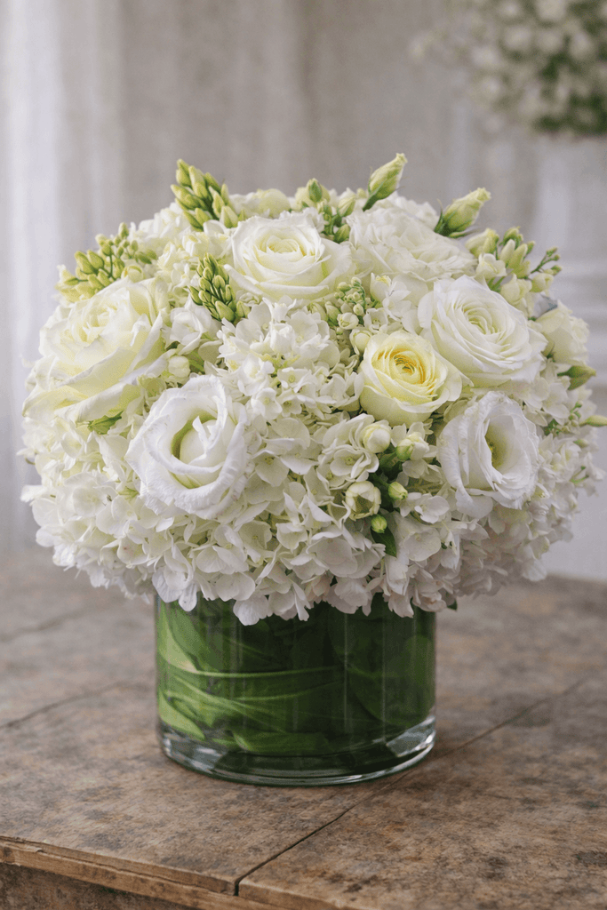White floral arrangement in a clear glass cylinder vase with white hydrangea, roses, lisianthus, and fresh seasonal blooms.
