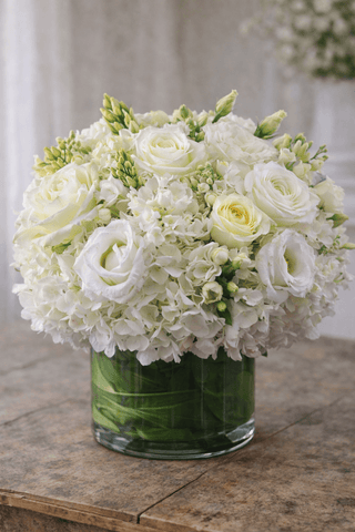 White floral arrangement in a clear glass cylinder vase with white hydrangea, roses, lisianthus, and fresh seasonal blooms.