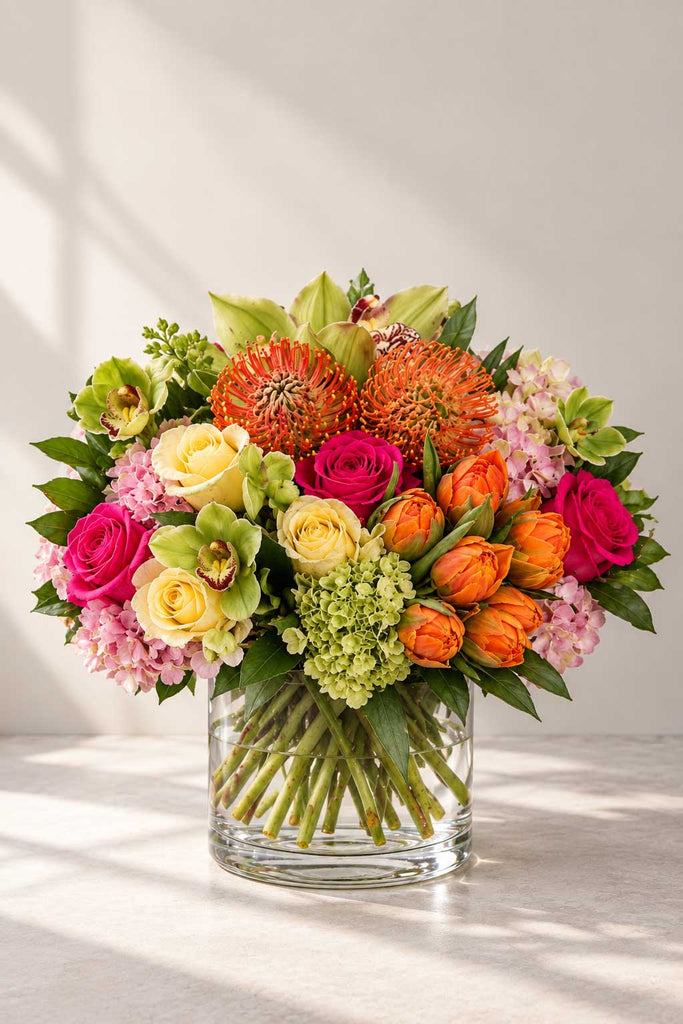 Sweet Surprise floral arrangement in a clear glass vase with roses, tulips, hydrangea, pincushion protea, cymbidium orchids, and seasonal blooms.