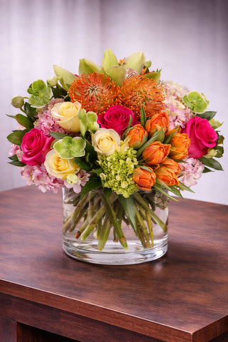 Sweet Surprise floral arrangement in a clear glass vase with roses, tulips, hydrangea, pincushion protea, cymbidium orchids, and seasonal blooms.