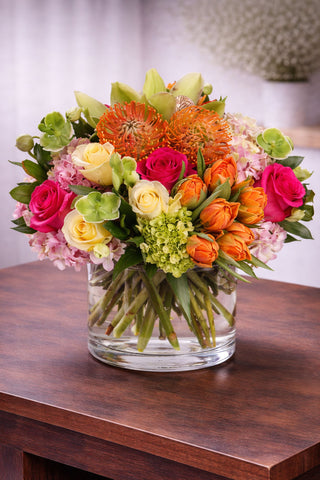Sweet Surprise floral arrangement in a clear glass vase with roses, tulips, hydrangea, pincushion protea, cymbidium orchids, and seasonal blooms.