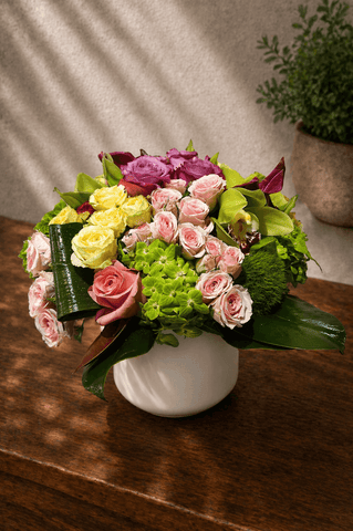 Pastel floral arrangement with pink and yellow roses, hydrangea, and seasonal blooms in a white ceramic vase on a neutral background