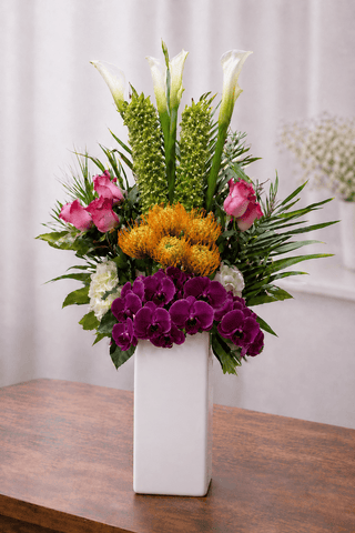 Tropical floral arrangement with purple orchids, white calla lilies, orange protea, pink roses, and greenery in a white rectangular vase.